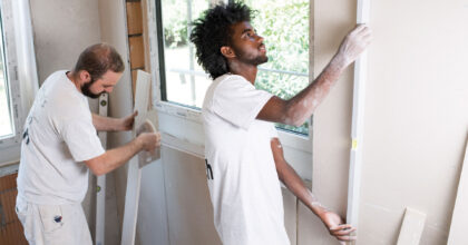 Ein Mann prüft mit einer Wasserwaage die vertikale Ausrichtung einer Wand, während ein zweiter Mann ein Holzstück vorbereitet.