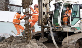 Auf der Baustelle verlassen zwei Männer einen Erdhaufen, während ein dritter Mann einen Erdbohrer bedient.