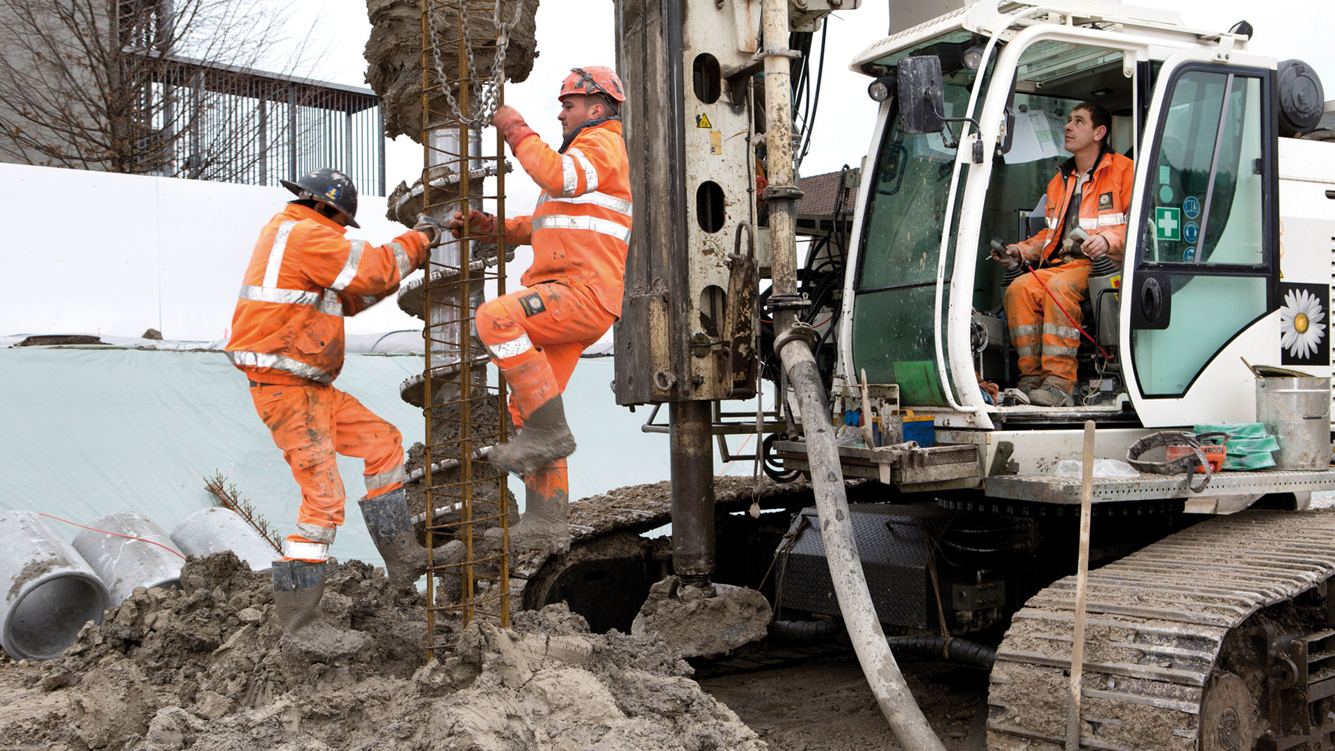 Auf der Baustelle verlassen zwei Männer einen Erdhaufen, während ein dritter Mann einen Erdbohrer bedient.