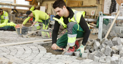 Ein Mann schlägt mit einem Pflästererhammer Pflastersteine in die Unterlage, während er auf einem niedrigen einbeinigen Stuhl sitzt.