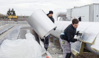 Ein Mann bringt Dämmung an Anlageteilen an, während ein zweiter Mann diese mit Blechen aus Leichtmetall abdeckt.