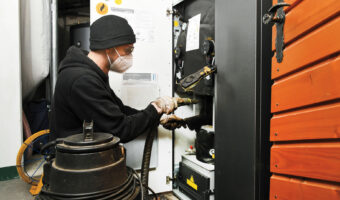 Ein Mann mit Mundschutz reinigt mit einem Staubsauger und einer Bürste eine wärmetechnische Anlage.