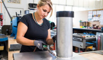 Eine Frau schweisst in einer Werkstatt ein Metallrohr an eine Metallplatte.