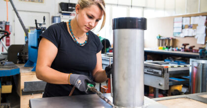 Eine Frau schweisst in einer Werkstatt ein Metallrohr an eine Metallplatte.