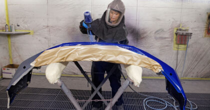 Eine Frau in einem Schutzanzug lackiert mit einer Spritzpistole die Stossstange eines Fahrzeugs.