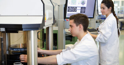 Ein Mann fertigt mit einer Fertigungsmaschine ein Glasprodukt, während eine Frau neben ihm einen Computer bedient.