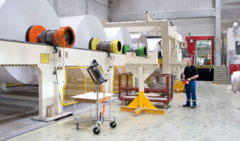 Ein Mann in einer Werkhalle kontrolliert riesige Papierrollen, die einen Prozess in einer Maschine durchlaufen.