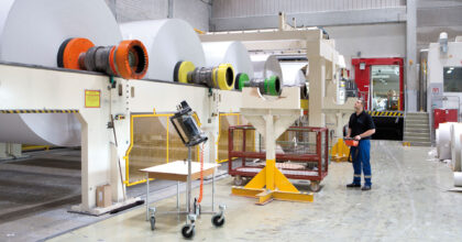 Ein Mann in einer Werkhalle kontrolliert riesige Papierrollen, die einen Prozess in einer Maschine durchlaufen.