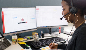 Eine Frau mit einem Headset schaut auf zwei Bildschirme, während sie sich Notizen macht.