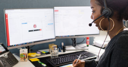 Eine Frau mit einem Headset schaut auf zwei Bildschirme, während sie sich Notizen macht.