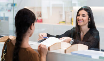 Eine Frau ist an einem Schalter im Gespräch mit einer Kundin und zeigt mit einem Kugelschreiber auf ein Dokument.