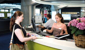 Eine Kundin wird bei der Ausleihe von Büchern von einer Frau an der Theke bedient.