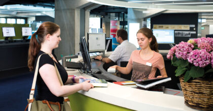 Eine Kundin wird bei der Ausleihe von Büchern von einer Frau an der Theke bedient.