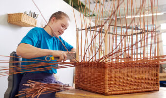 Eine Frau sitzt an einem Tisch und flechtet an einem Korb.