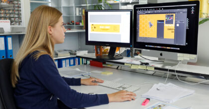 Eine Frau sitzt vor zwei Bildschirmen und layoutet eine Etikette für ein Honigglas.