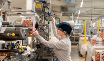 In einer Werkhalle spannt eine Frau mit Schutzbrille und Ohrstöpsel eine Rolle einer Verpackung in eine Maschine.