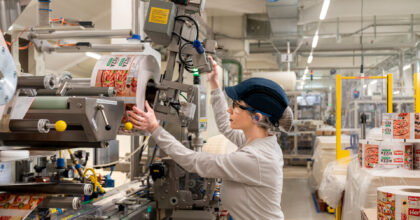 In einer Werkhalle spannt eine Frau mit Schutzbrille und Ohrstöpsel eine Rolle einer Verpackung in eine Maschine.