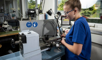 Eine Frau mit Schutzbrille steht vor einer Maschine und stellt ein Metallteil her.