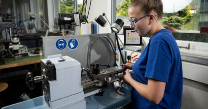 Eine Frau mit Schutzbrille steht vor einer Maschine und stellt ein Metallteil her.