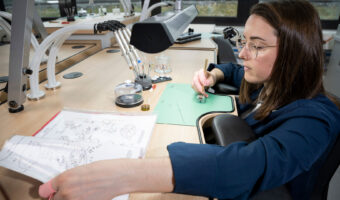 Eine Frau montiert nach einem Plan ein Uhrwerk.