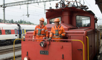 Eine Frau und ein Mann stehen auf einer Rangierlok und sehen auf die Geleise.
