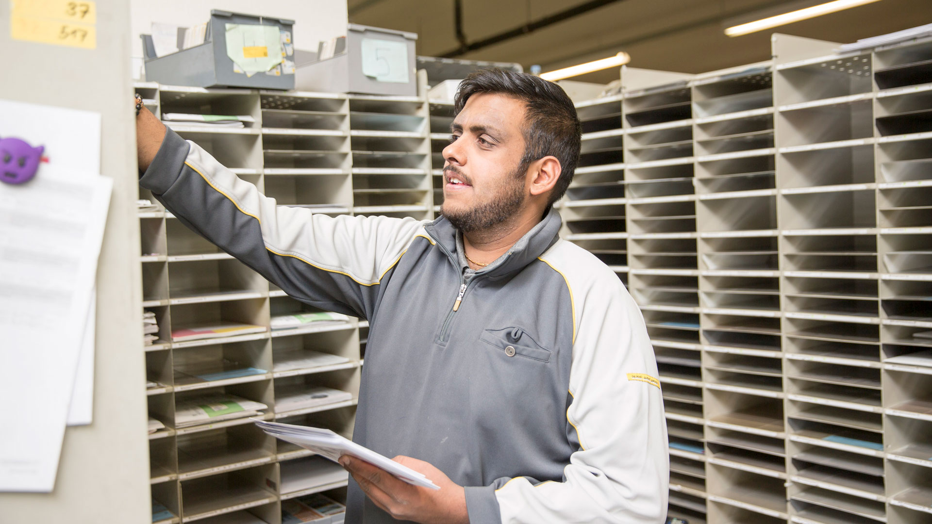 Ein Mann steht vor einer Wand mit vielen Fächern und sortiert Briefe ein.