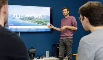 Ein Mann steht vor einem grossen Bildschirm und erklärt zwei weiteren Männern die Präsentation die zu sehen ist.