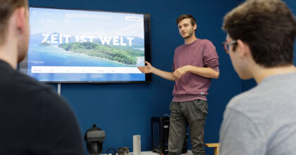 Ein Mann steht vor einem grossen Bildschirm und erklärt zwei weiteren Männern die Präsentation die zu sehen ist.