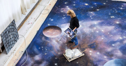 Eine Frau die in Socken auf einem grossen Bild am Boden steht, hält in der einen Hand die verkleinerte Vorlage des Bildes und in der anderen Hand einen Pinsel mit sehr langem Stiel und malt.