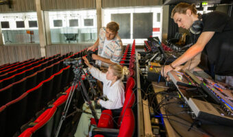 In einem Saal installieren eine Frau und ein Mann eine Kamera. Im Hintergrund richtet ein weiterer Mann ein Mischpult ein.