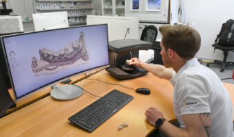 Ein Zahntechniker hält eine Zahnprothese unter einen Scanner um sie an einem Bildschirm zu betrachten.