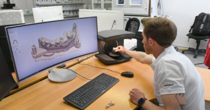 Ein Zahntechniker hält eine Zahnprothese unter einen Scanner um sie an einem Bildschirm zu betrachten.