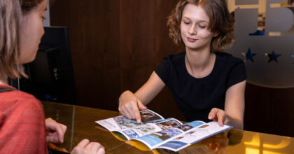 Eine Frau hinter einem Hoteldesk erklärt einem weiblichen Gast, anhand eines Planes, den Weg zu einer Sehenswürdigkeit.