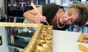 Eine Frau trägt mit einem Pinsel Goldfolie auf einen verschnörkelten Spiegelrahmen auf.