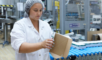 Eine Frau steht an einem Fliessband und prüft eine Kartonschachtel.