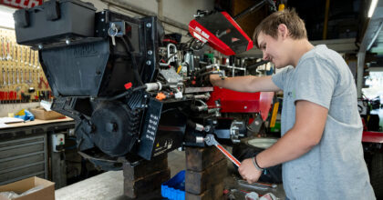 Ein lächelnder junger Mann hantiert am Motor eines Balkenmähers.