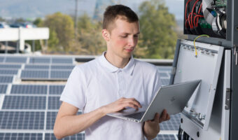 Ein junger Mann steht mit einem Laptop in den Händen auf einem Dach mit Solarpanels und prüft die Funktion.