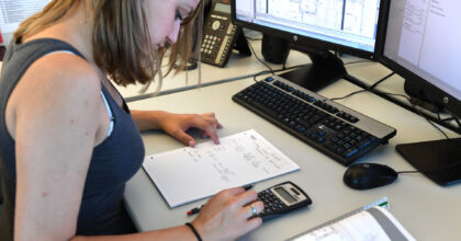 Eine junge Frau sitzt an einem Arbeitsplatz und rechnet mit einem Taschenrechner Masse aus.