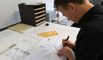 Ein Mann sitzt an einem Arbeitstisch und zeichnet von Hand mit Hilfe eines Geodreiecks auf seinem Arbeitsblatt.