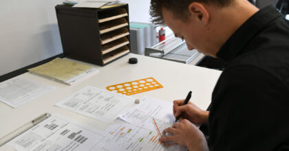 Ein Mann sitzt an einem Arbeitstisch und zeichnet von Hand mit Hilfe eines Geodreiecks auf seinem Arbeitsblatt.