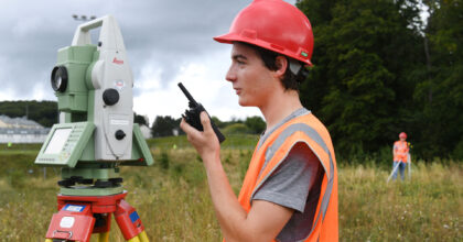 Ein junger Mann steht auf einer Wiese hinter seinem Messgerät und spricht in ein Funkgerät.
