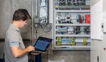 Ein Mann mit einem Laptop in den Händen steht vor einer geöffneten Kälteanlage und prüft die Funktion.